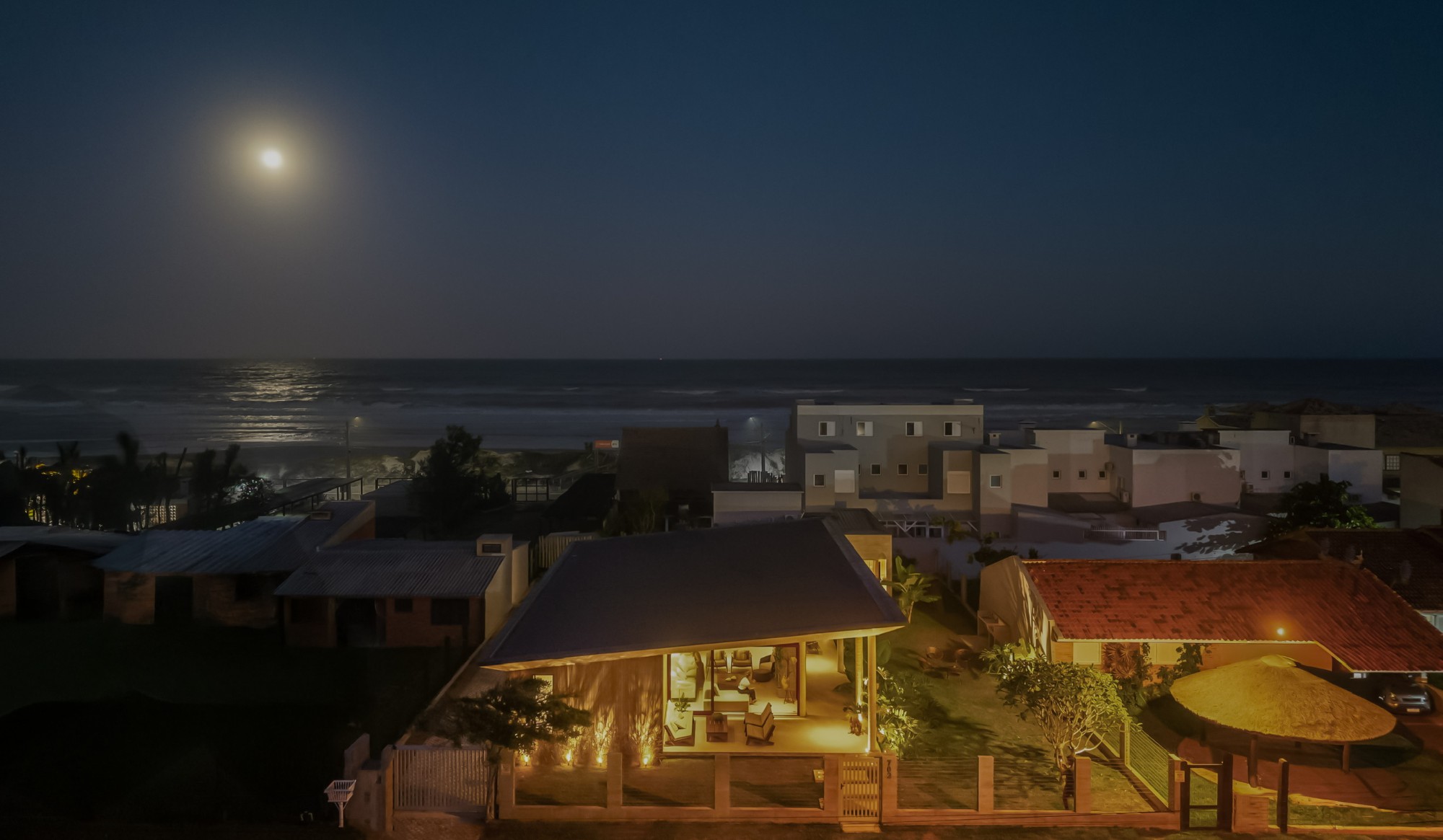 Vista aérea noturna da casa, com o mar e a lua ao fundo